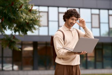 Handsome young man smiles while using his laptop outside a contemporary building in the sunlight.