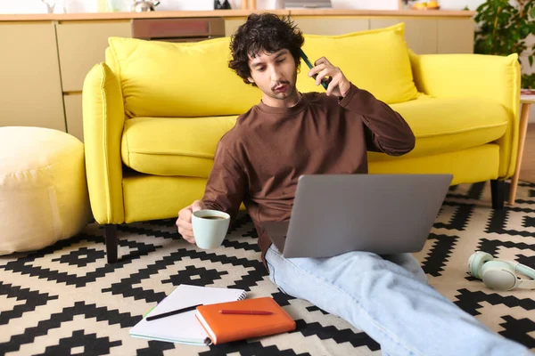 A handsome young man enjoys a work session at home, talking on the phone and sipping coffee.