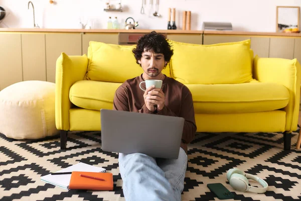 A handsome young man relaxes on the floor with coffee, focused on his laptop in a stylish room.