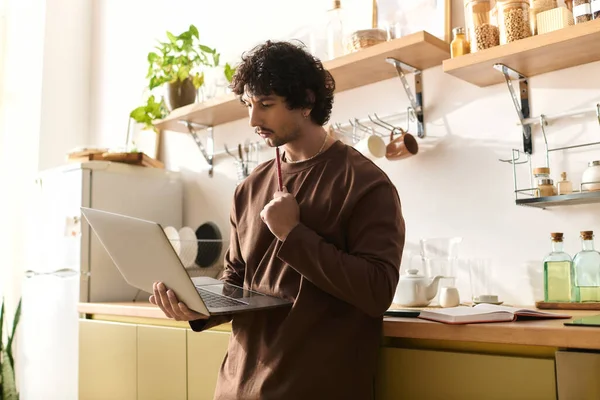 A young man stands in a modern kitchen, engaged with his laptop while contemplating ideas.