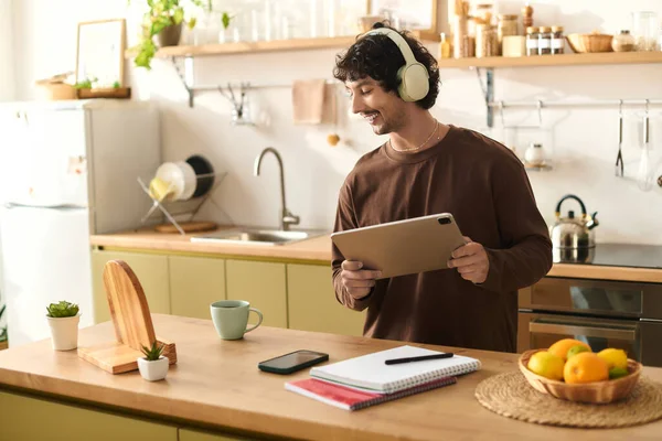 Young man with headphones smiles while using a tablet in his warm kitchen environment.