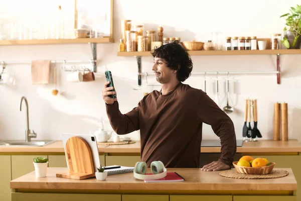 Handsome young man stands in a modern kitchen, smiling while looking at his smartphone.
