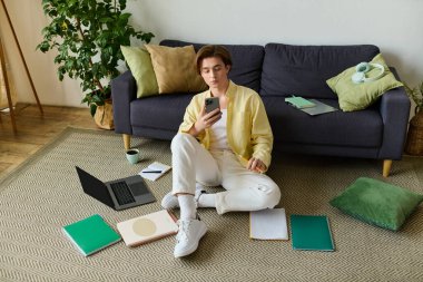 Şık giyinmiş yakışıklı genç adam rahat bir yaşam alanının ortasındaki akıllı telefonuna odaklanıyor..