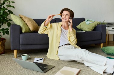 Tarz sahibi genç adam yerde otururken gülümsüyor, evde dizüstü bilgisayar ve telefonla çalışıyor..