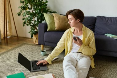 Genç adam yerde oturuyor, rahat bir yerde dizüstü bilgisayar ve telefon kullanıyor..