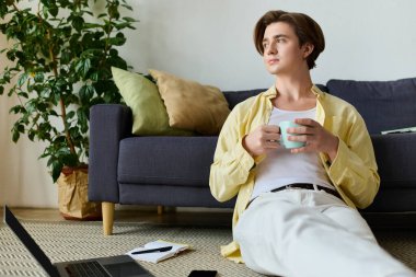 Yakışıklı genç adam elinde kahve fincanıyla yerde dinleniyor. Etrafı şık dekorlar ve bitkilerle çevrili..