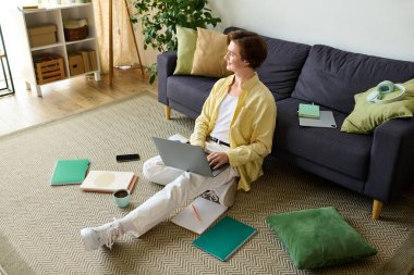 Genç, modaya uygun bir adam yerde oturuyor, parlak bir yaşam alanında dizüstü bilgisayarına odaklanmış..