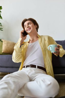 Şık bir genç adam yerde oturup çay yudumlarken mutlu bir şekilde telefonla konuşuyor..