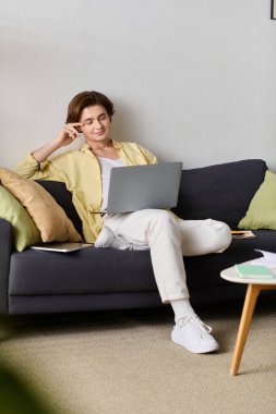 Genç, modaya uygun bir adam, modern bir oturma odasında dizüstü bilgisayarıyla rahat bir şekilde oturur..