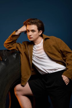 Yakışıklı genç adam kendine güvenerek koyu mavi bir zemine karşı poz veriyor, moda kıyafetinin altını çiziyor..