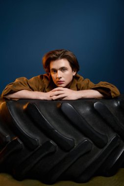 Genç adam büyük bir lastiğe yaslanıyor, sanatsal bir iç mekanda moda bir kıyafet sergiliyor..