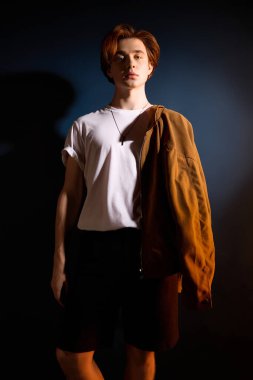 Yakışıklı genç adam gündelik bir duruş sergiliyor, hafif ışıklandırmayla modaya uygun kıyafetlerini sergiliyor.