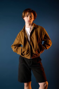 Modaya uygun kahverengi ceketli ve siyah şortlu yakışıklı genç adam mavi bir duvarın yanında poz veriyor..