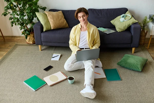 Genç, yakışıklı bir adam yerde oturuyor, neşeli bir şekilde dizüstü bilgisayarına odaklanmış..