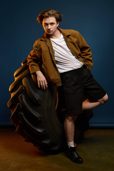 Fashionable young man poses beside an oversized tire, showcasing his trendy style and confidence.