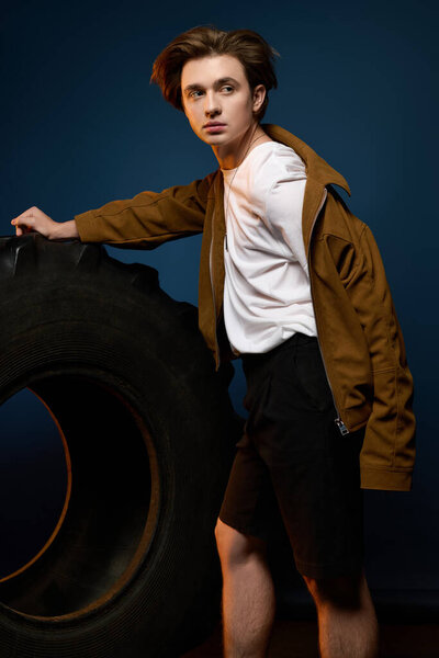 A stylish young man poses confidently by a large tire, flaunting his trendy outfit.