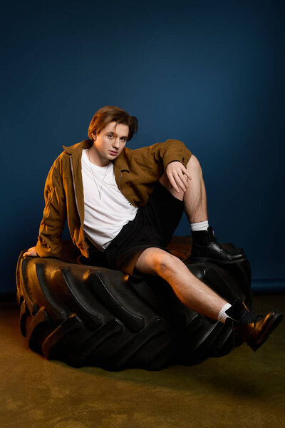 Handsome young man showcases stylish outfit while relaxing on a massive tire against a blue backdrop