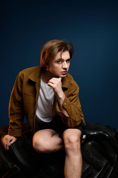 A handsome young man sits thoughtfully on a tire while showcasing his trendy outfit in a studio.