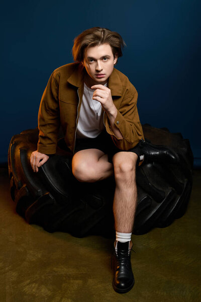 A stylish young man sits on a tire, showcasing his trendy outfit and relaxed demeanor indoors.