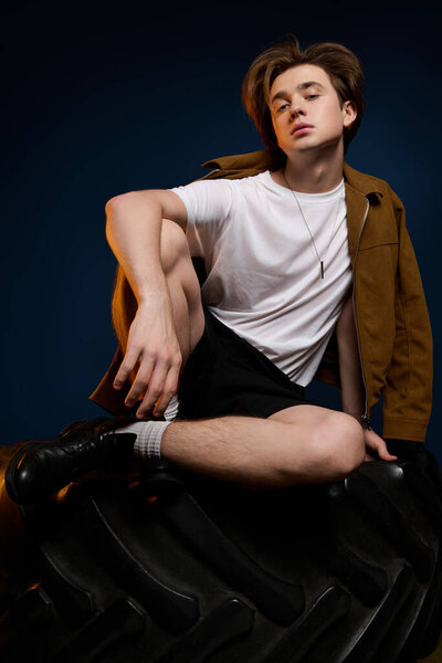 Stylish young man with confident posture sits on a giant tire, showcasing modern fashion.