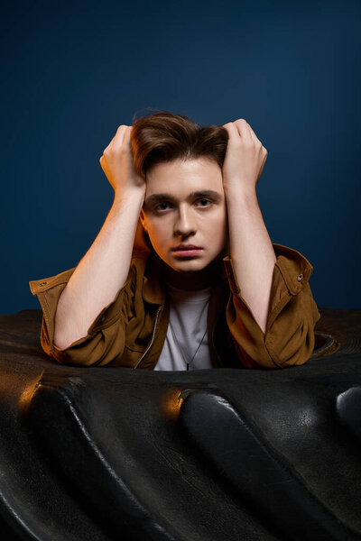 A young, fashionable man expresses emotion with his hands on his head, showcasing a striking look.