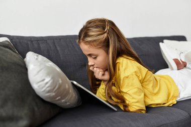 Genç bir kız kanepede dinleniyor, tabletine dalmış, alfa çocuk.