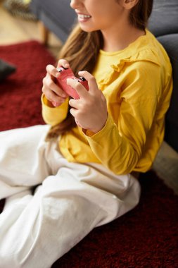 Genç bir kız evdeki yumuşak halıya oturmuş, günlük kıyafetleriyle video oyunları oynuyor.