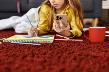 Gen alfa kızı evde vakit geçirmeyi sever, defterini çizer ve rahat bir halının üzerinde telefonunu kontrol eder..