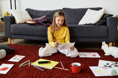 Sıradan elbiseli bir kız renkli çizimler ve sanat malzemeleriyle çevrili yerde oturuyor..