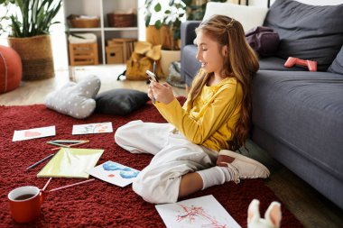 Neşeli bir kız evdeki renkli çizimleri ve sanat aletleriyle çevrili rahat bir halıya oturur..