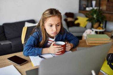 Bir kız evinde, elinde fincanıyla rahatlıyor, ödevine ve ekranına odaklanıyor..