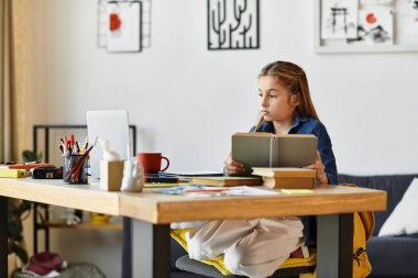 Evde çalışma materyalleriyle çevrelenmiş bir kız, rahatça bir masada oturup kitap okuyor..