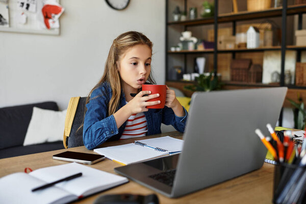 Молодая девушка сидит за столом, пьет из красной кружки, фокусируясь на своем ноутбуке..