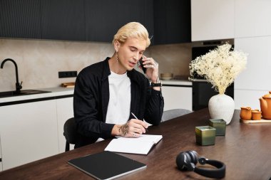 Yakışıklı genç adam telefonda konuşuyor ve modern bir mutfak masasına notlar yazıyor..