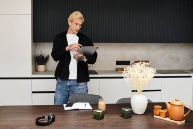Yakışıklı genç bir adam şık bir dairede, bir tablet üzerinde düşünceli bir şekilde çalışıyor ve tarzını gösteriyor..