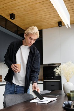 Yakışıklı genç adam bir içki tutuyor ve telefonunu dairesindeki ahşap bir masaya yerleştiriyor..