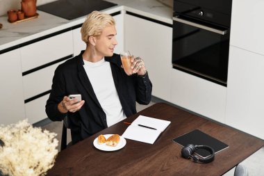 Yakışıklı bir adam modern bir masada oturmuş, içkisini yudumluyor ve telefonunu kontrol ediyor, rahat ve odaklanmış..