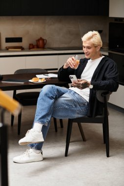 Yakışıklı genç bir adam modern bir dairede rahatça oturur, içkisini yudumlar ve telefonunu kontrol eder..