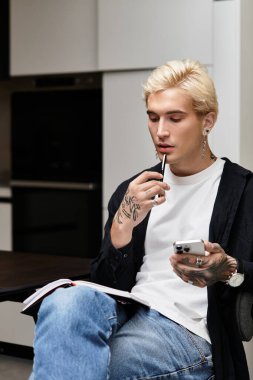 Düşünceli bir genç adam şık bir dairede oturuyor, elinde kalem ve telefon var..