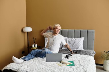 Neşeli genç bir adam rahat bir yatakta oturur, gülümser ve modern bir dairede dizüstü bilgisayarıyla meşgul olur..