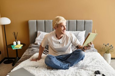 Genç adam rahat bir yatakta oturuyor, bir kitaba dalmış, modern bir apartman atmosferiyle çevrili..