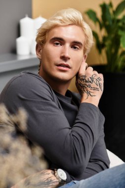 Genç bir adam zarif bir dairede oturuyor, dövmelerini ve cazibesini sergiliyor..