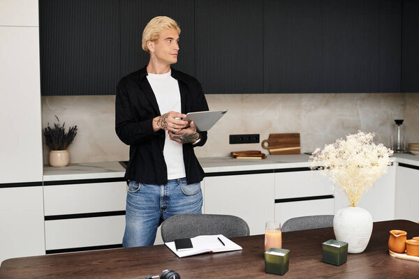 A handsome young man stands in a chic apartment, engaged in creative work with a tablet in hand.