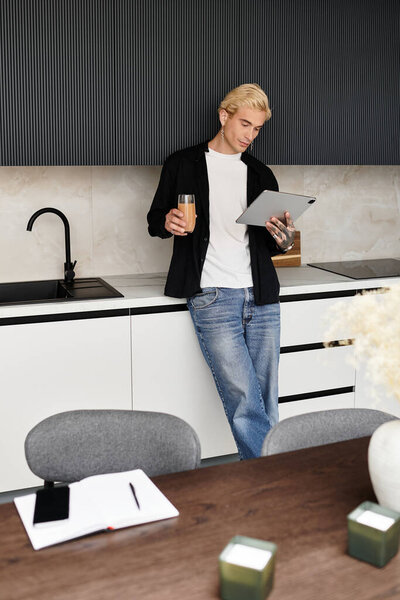 A young and handsome man stands in a modern kitchen, sipping a drink and using a tablet.