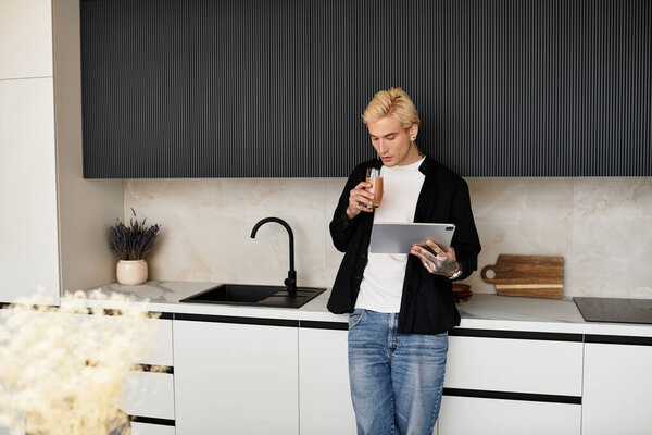 Handsome young man relaxes in a stylish kitchen, sipping a drink and browsing on his tablet.