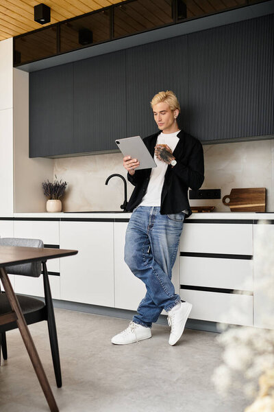 A young man with blond hair casually reads on his tablet in a stylish kitchen.