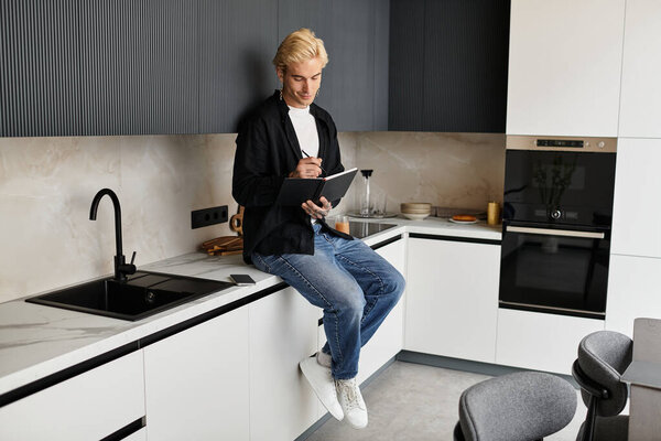 A young man sits on a countertop in a contemporary kitchen, jotting down thoughts with a pen.