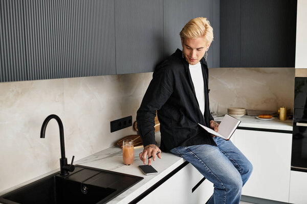 Stylish young man sits on a kitchen counter, sipping a drink and jotting down ideas in a notebook.
