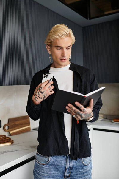 Красивый молодой человек с татуировками сфокусирован на блокноте в современной кухне.
