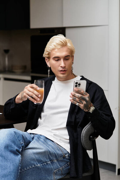 Стильный молодой человек удобно сидит в шикарной квартире, потягивая напиток и пользуясь телефоном.